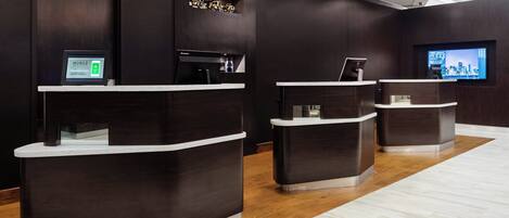 A reception area with three dark wooden reception desks, a digital screen, and a television displaying a cityscape.