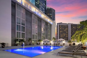 Un espace piscine d’hôtel avec des chaises longues et des parasols, entouré de grands immeubles et surplombant un ciel au coucher du soleil.