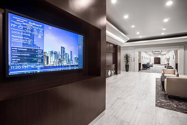 Vestíbulo de hotel con una gran pantalla digital que muestra eventos locales y el pronóstico del tiempo, una recepción y un área de descanso con vista a la ciudad.