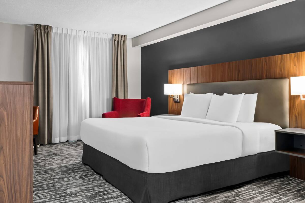 A hotel room with a large bed, a red chair, a wooden wardrobe, and a nightstand with a lamp.
