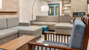 A modern living room with a sectional sofa, a wooden coffee table, and a blue ottoman. There is a wooden chair with a patterned cushion, a floor lamp, and a bookshelf with books. A television is mounted on the wall.