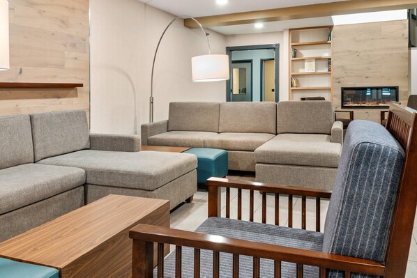 A modern living room with a sectional sofa, a wooden coffee table, and a blue ottoman. There is a wooden chair with a patterned cushion, a floor lamp, and a bookshelf with books. A television is mounted on the wall.