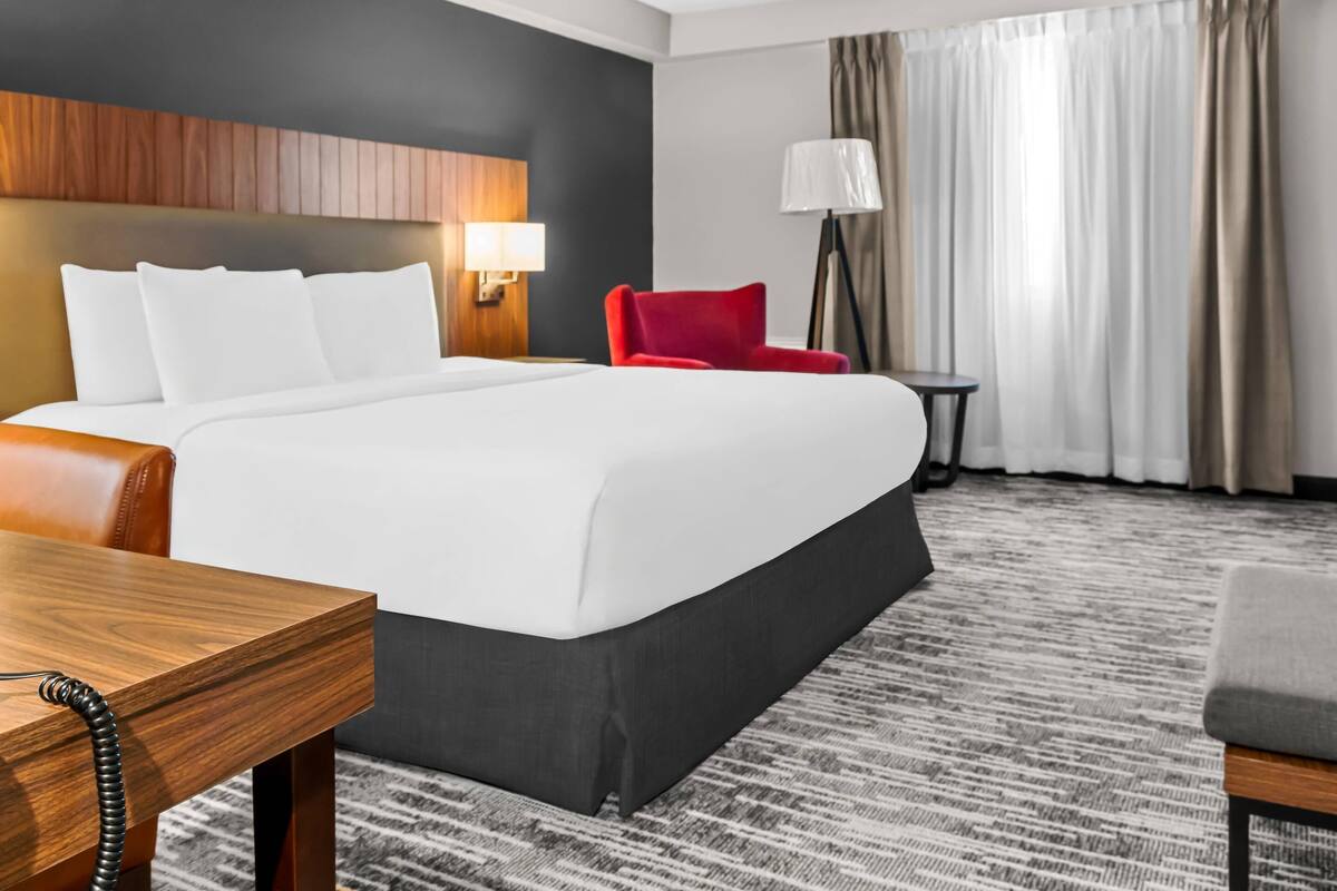 A hotel room with a large bed, a red chair, a small table, and a lamp.