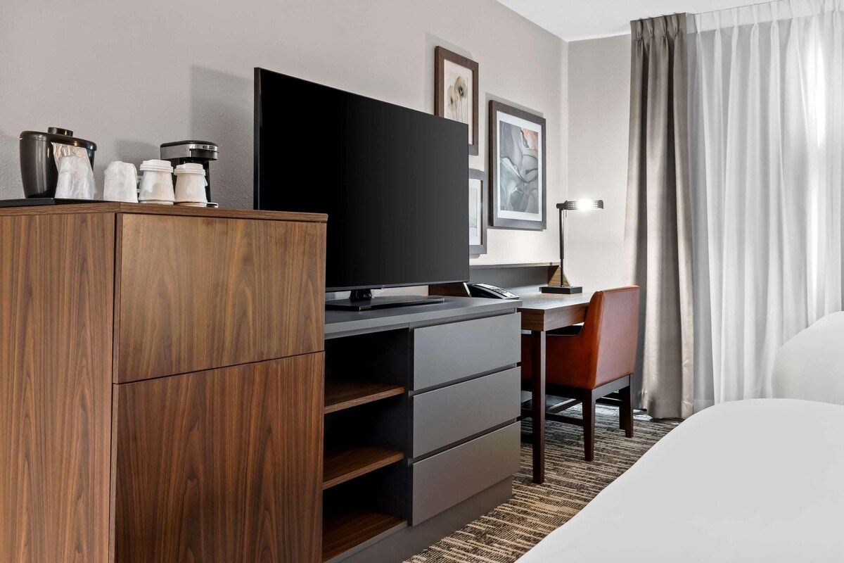 A modern hotel room with a wooden wardrobe, a flat-screen TV, a desk with a chair, and a bed.
