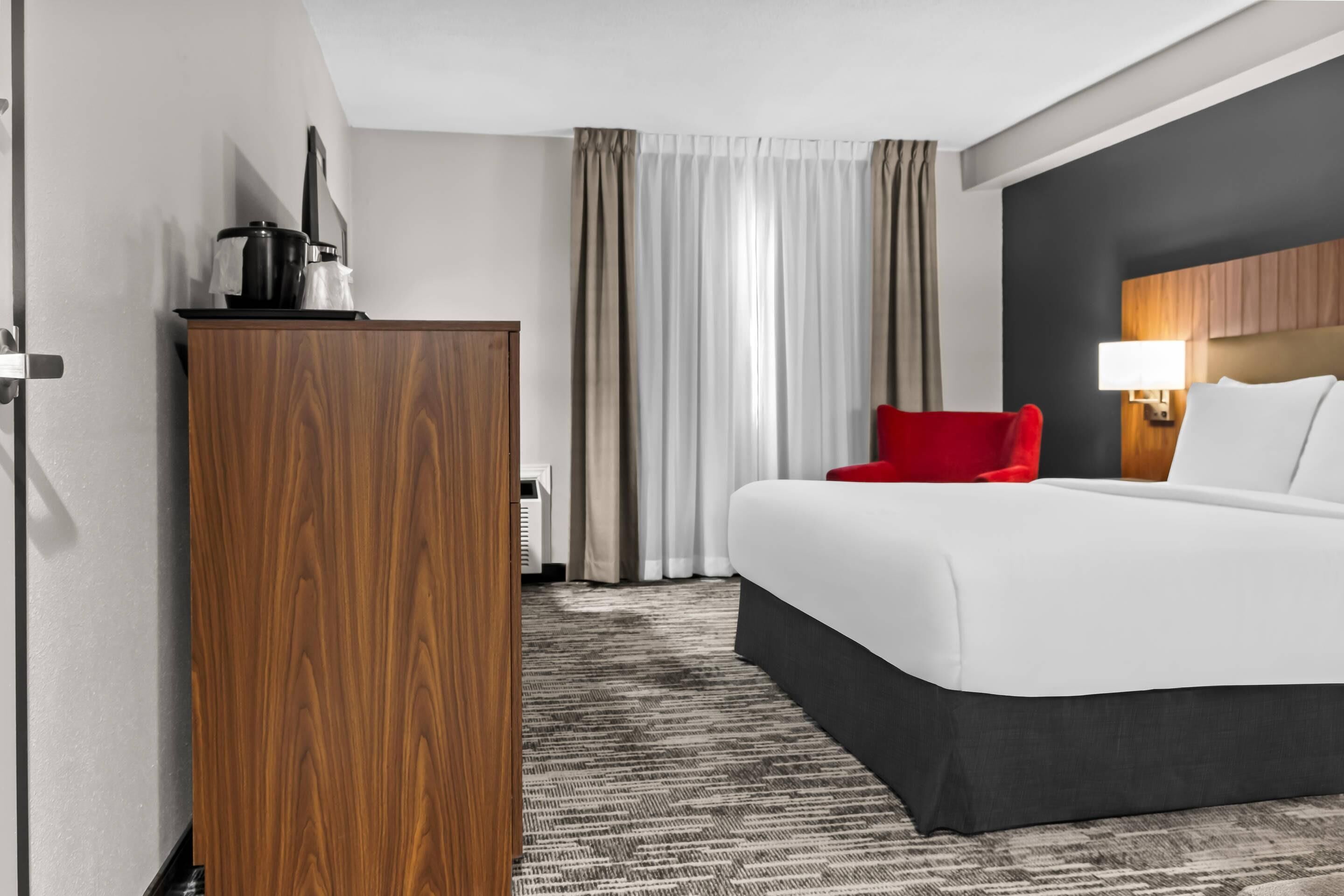 A hotel room with a large bed, a red chair, a wooden wardrobe, a desk with a lamp, and a view of the outside through curtains.