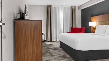 A hotel room with a large bed, a red chair, a wooden wardrobe, a desk with a lamp, and a view of the outside through curtains.