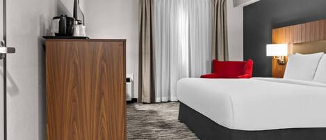 Una habitación de hotel con una cama grande, una silla roja, un armario de madera, un escritorio con una lámpara y vistas al exterior a través de cortinas.
