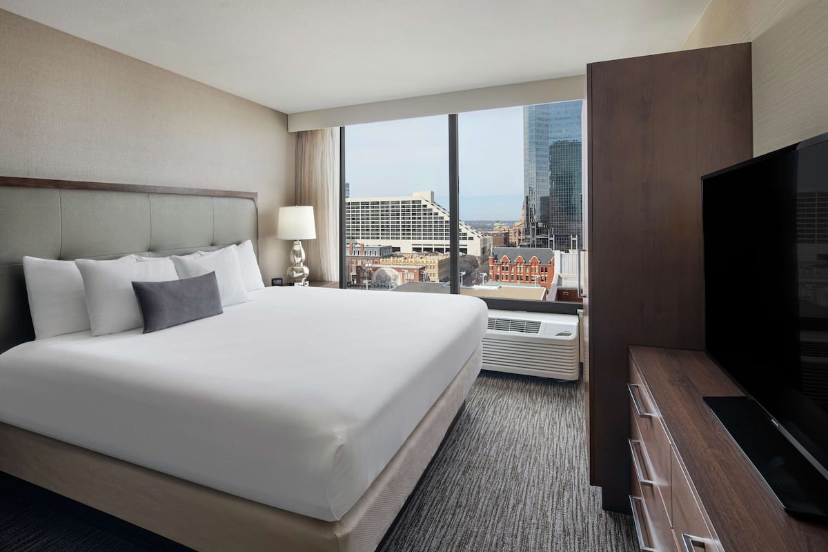 A hotel room with a large bed, a flat-screen TV, a desk, and a view of the cityscape.
