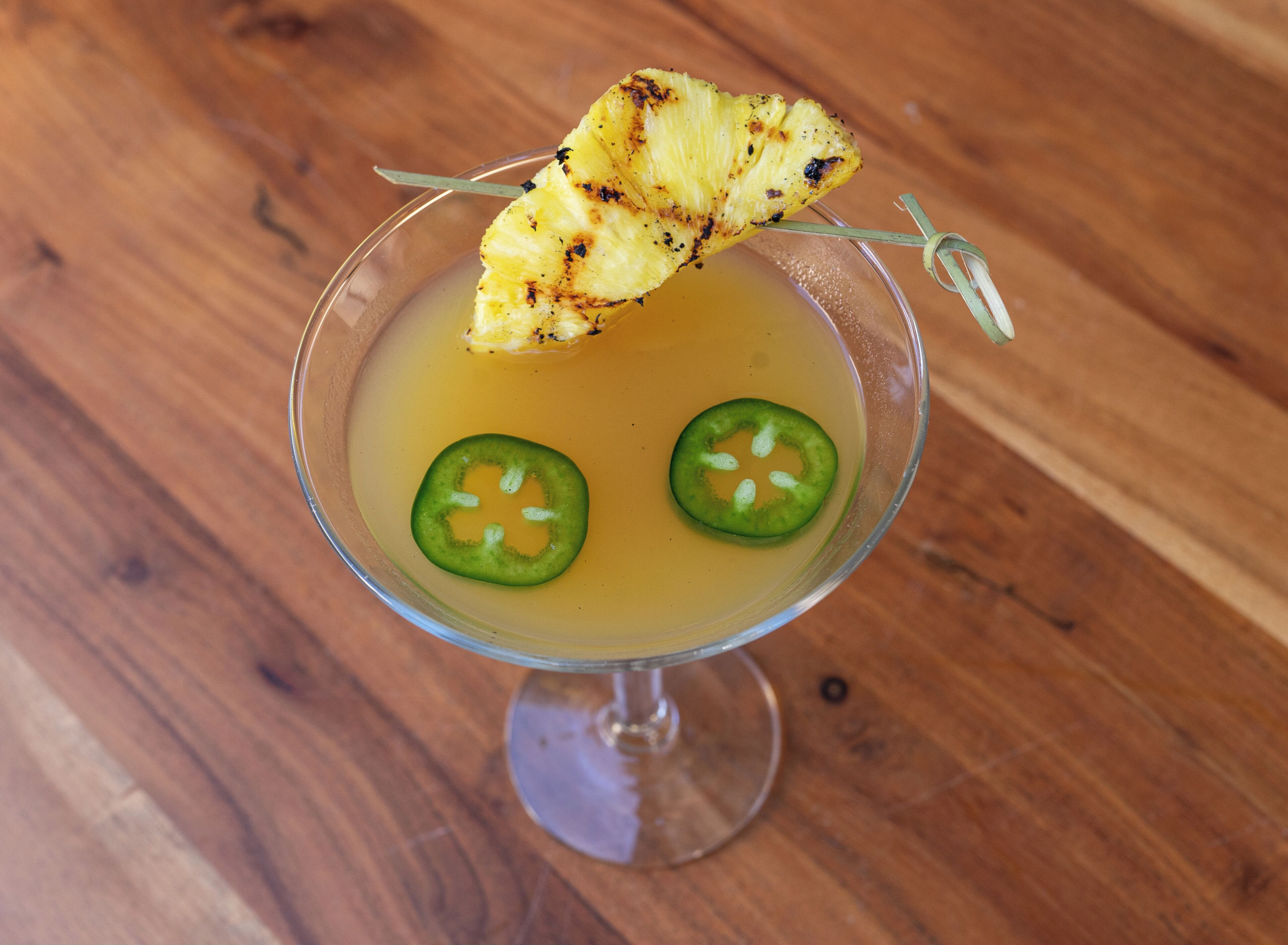 Een cocktail met een ananasstukje en jalapeño-schijfjes op een houten tafel.