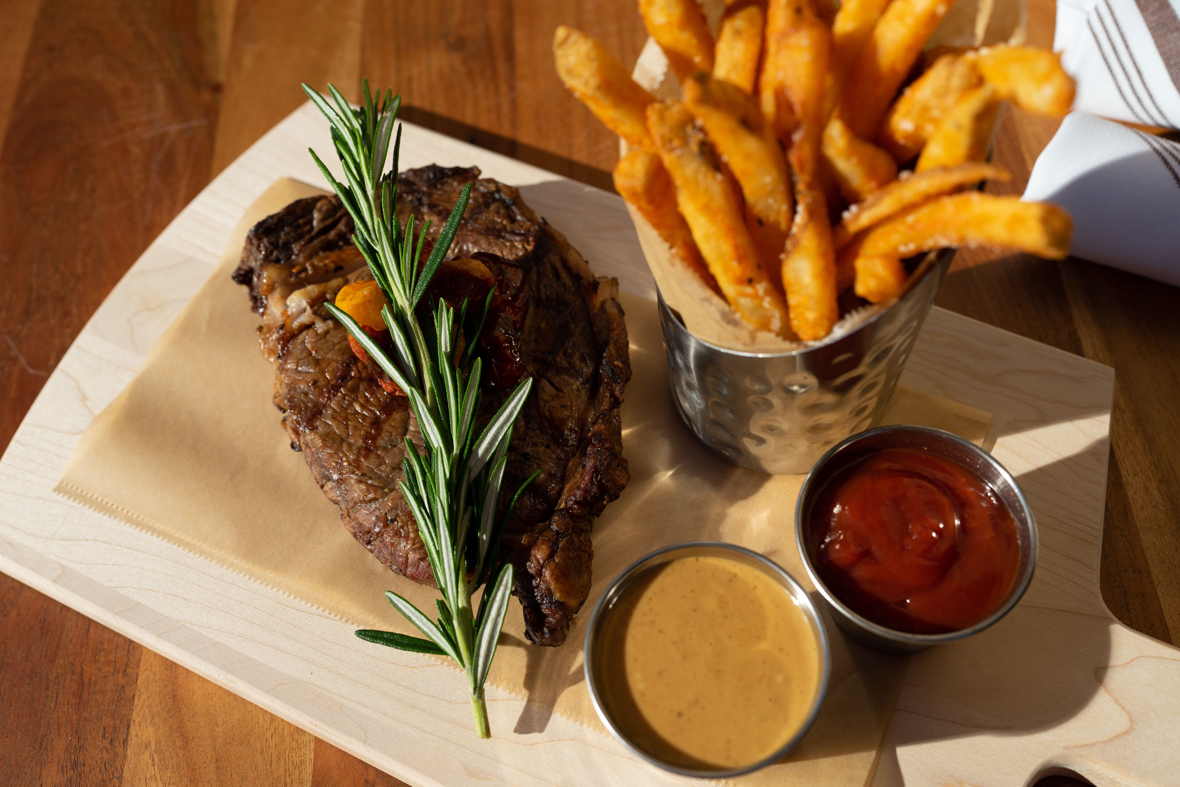 A steak with rosemary garnish, a side of fries, and two dipping sauces on a wooden table.