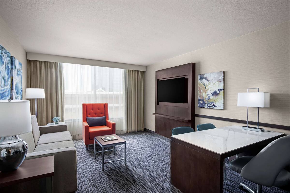 A hotel room with a sofa, armchair, desk, TV, and artwork on the wall.