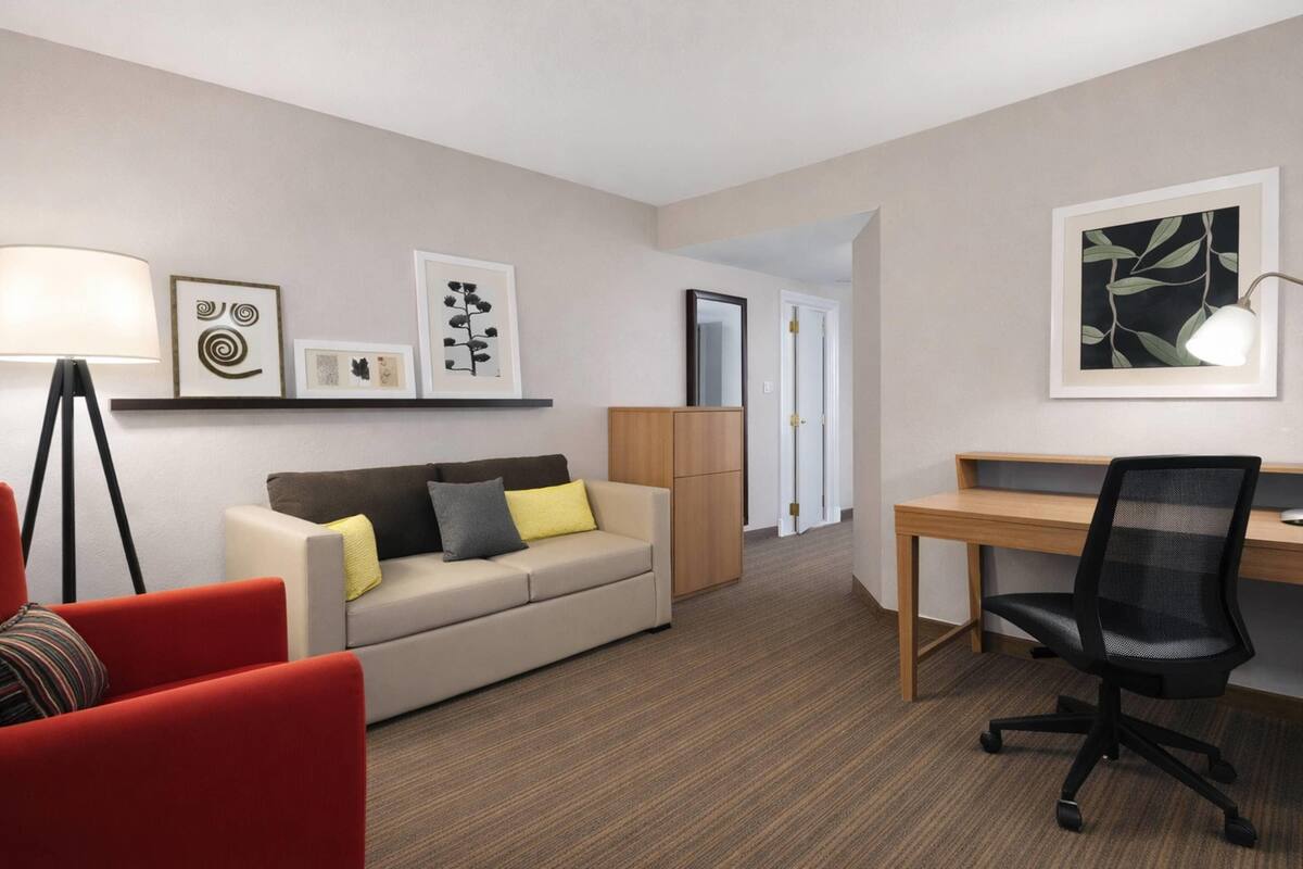 A hotel room with a sofa, a desk, a chair, a lamp, and artwork on the walls.