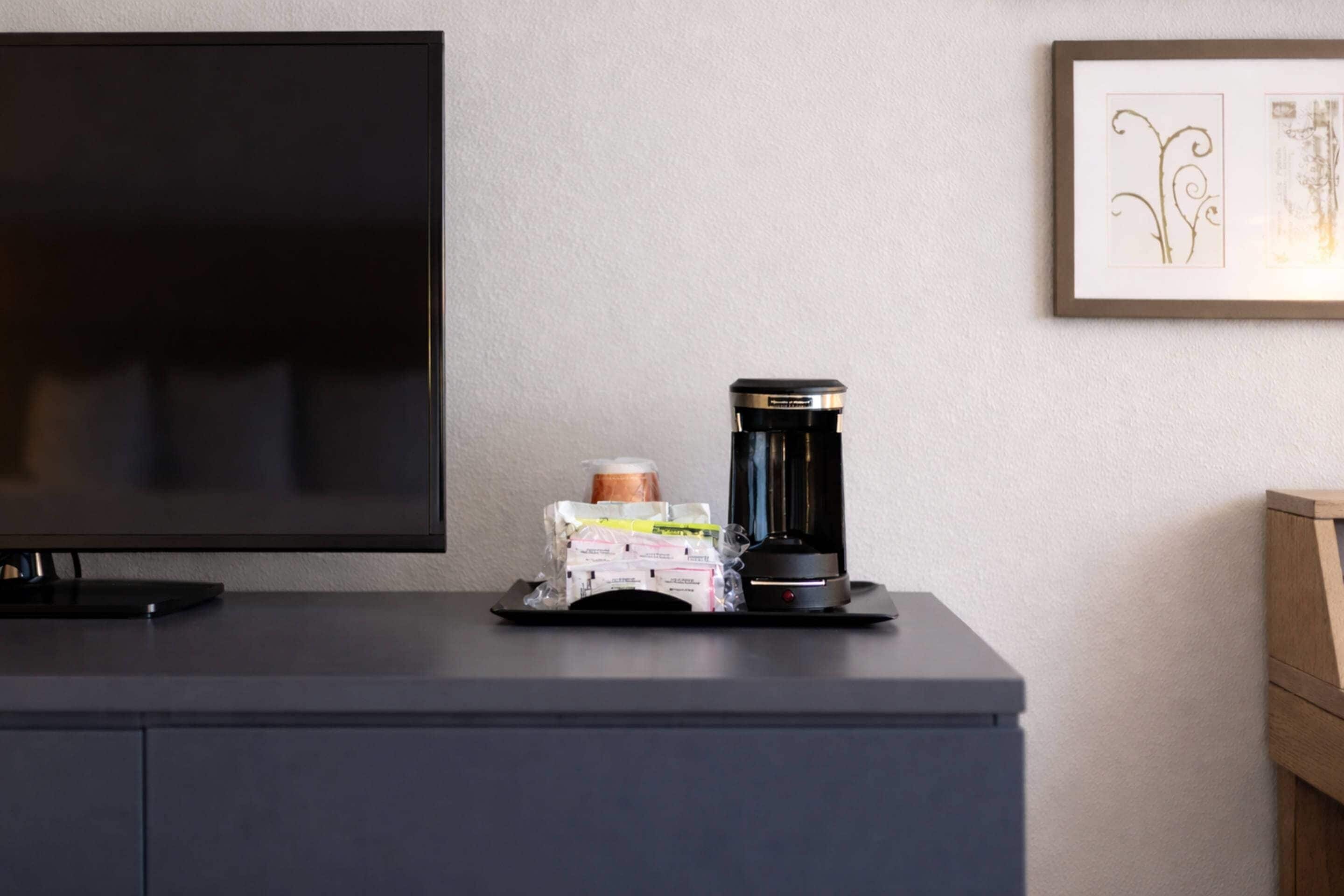 A flat-screen TV on a dark-colored cabinet, a coffee maker, and a framed artwork on the wall.