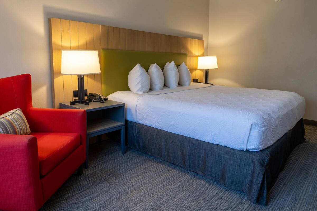 A hotel room with a bed, two bedside lamps, a red armchair, a telephone, and a small table.