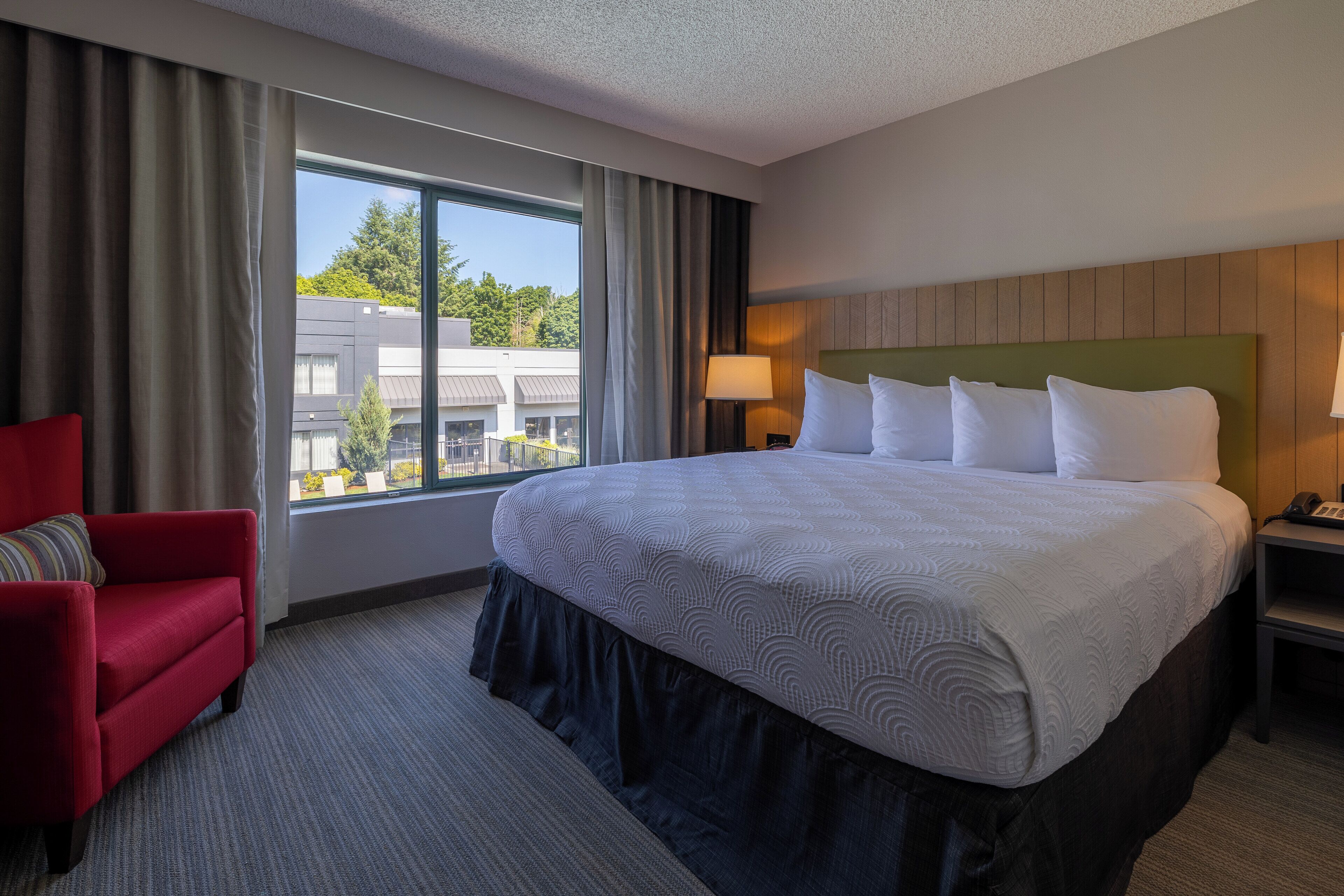 Habitación de hotel con una cama grande, un sillón rojo, una mesita de noche y una ventana con vistas a edificios y árboles.