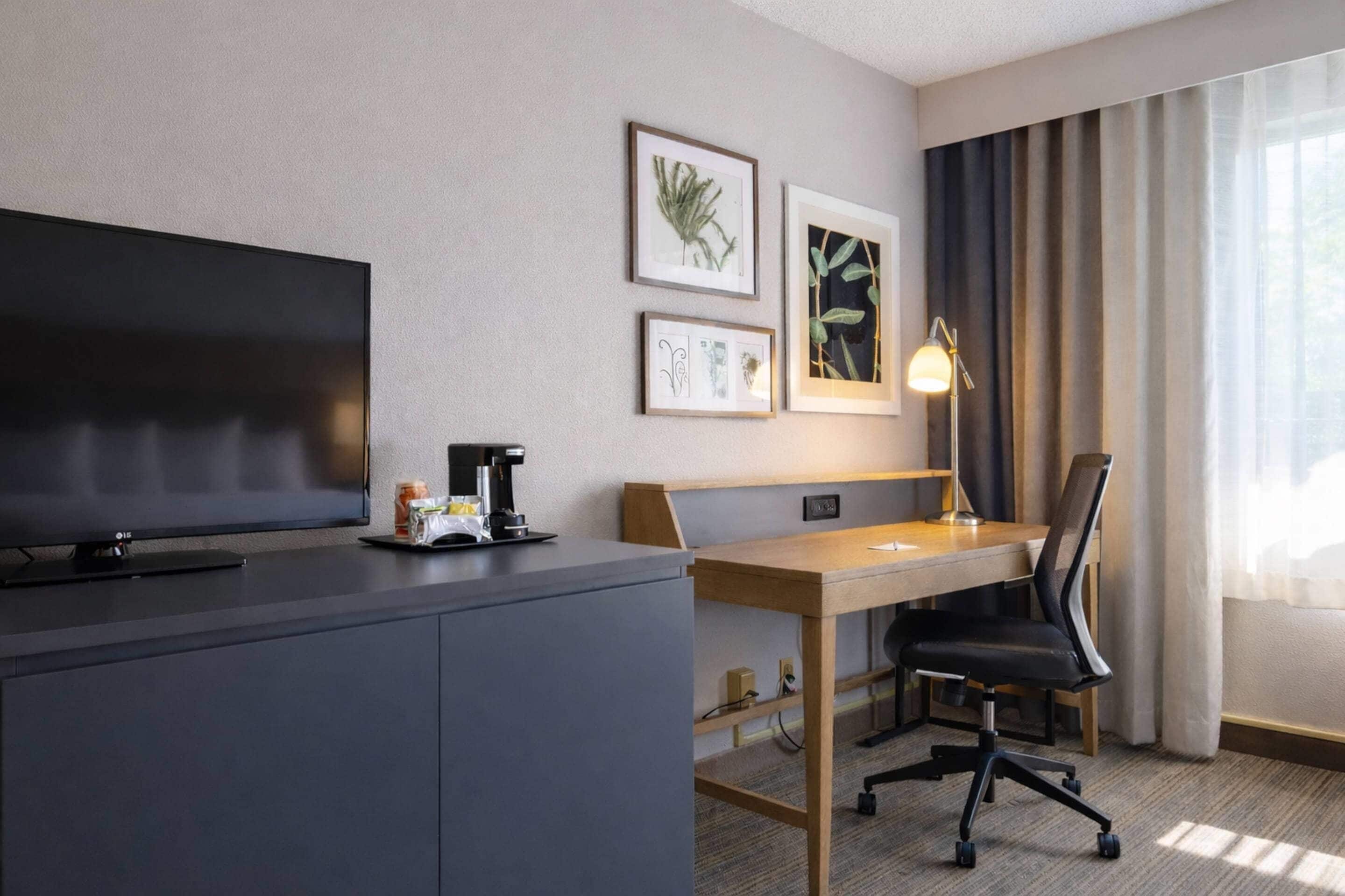 Habitación de hotel con televisor de pantalla plana, escritorio con silla, lámpara e impresiones botánicas enmarcadas en la pared.