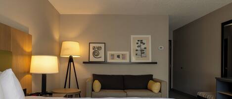 A hotel room with a bed, a bedside table, a lamp, a sofa, and wall art.