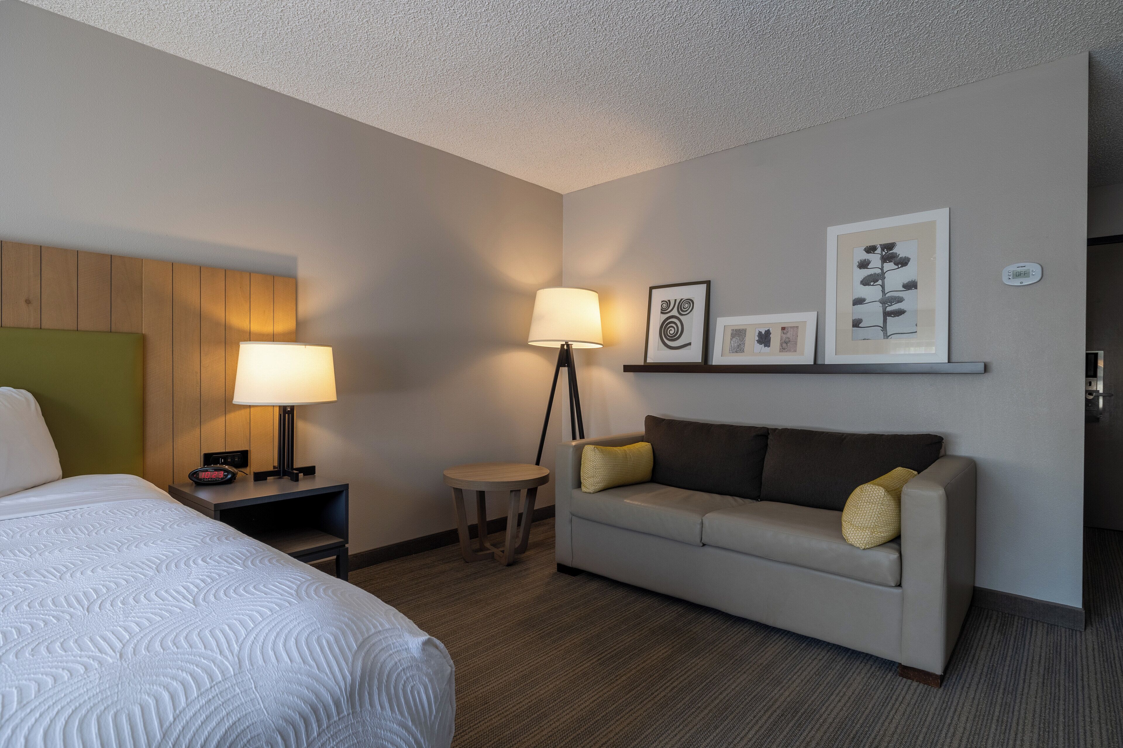 A hotel room with a bed, a sofa, a small table, a lamp, and wall art.