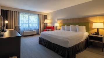 A hotel room with a large bed, two bedside tables with lamps, a desk with a chair, a red armchair, and a large window with curtains.