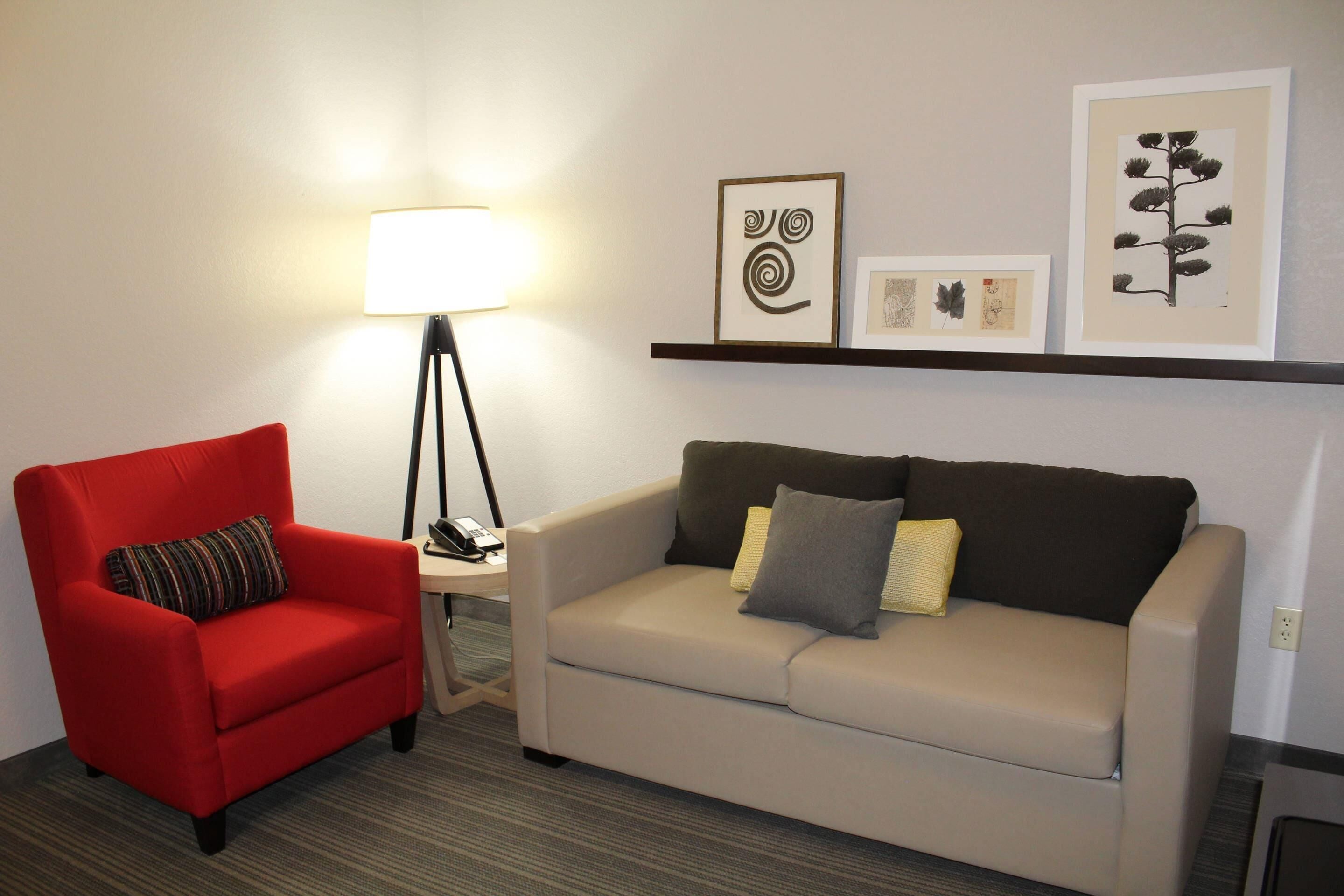 A room with a red armchair, a beige sofa, a floor lamp, and framed artwork on the wall.