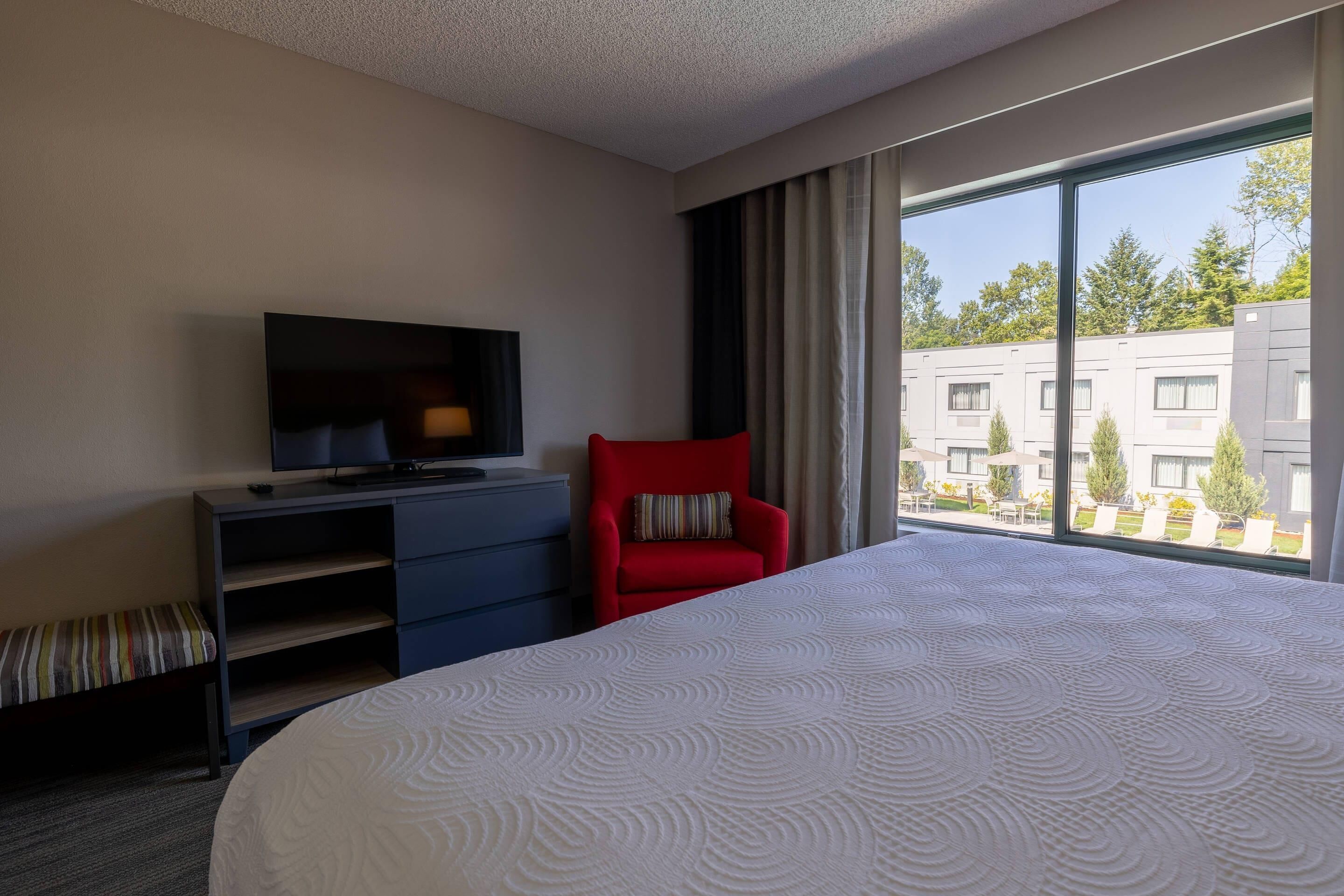A hotel room with a bed, a TV on a desk, a red chair, and a large window with a view of buildings.