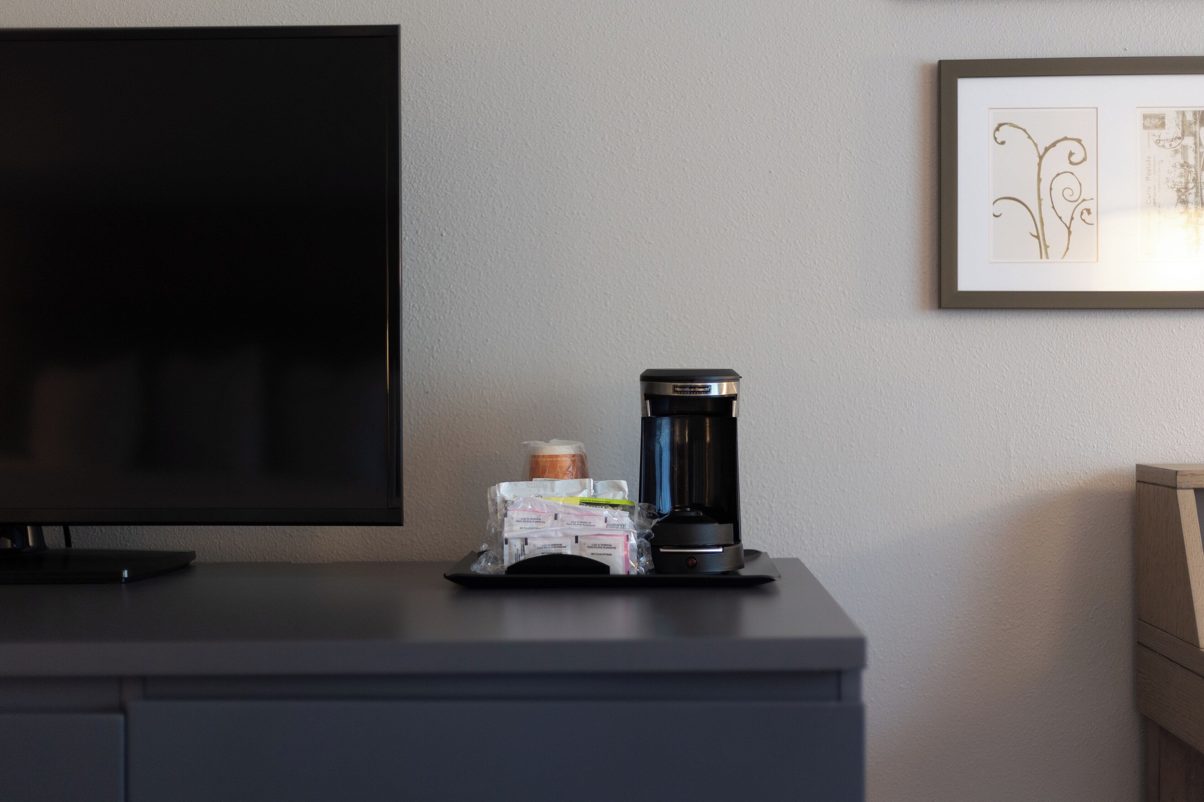 A flat-screen TV, a coffee maker, and a framed picture on a desk.