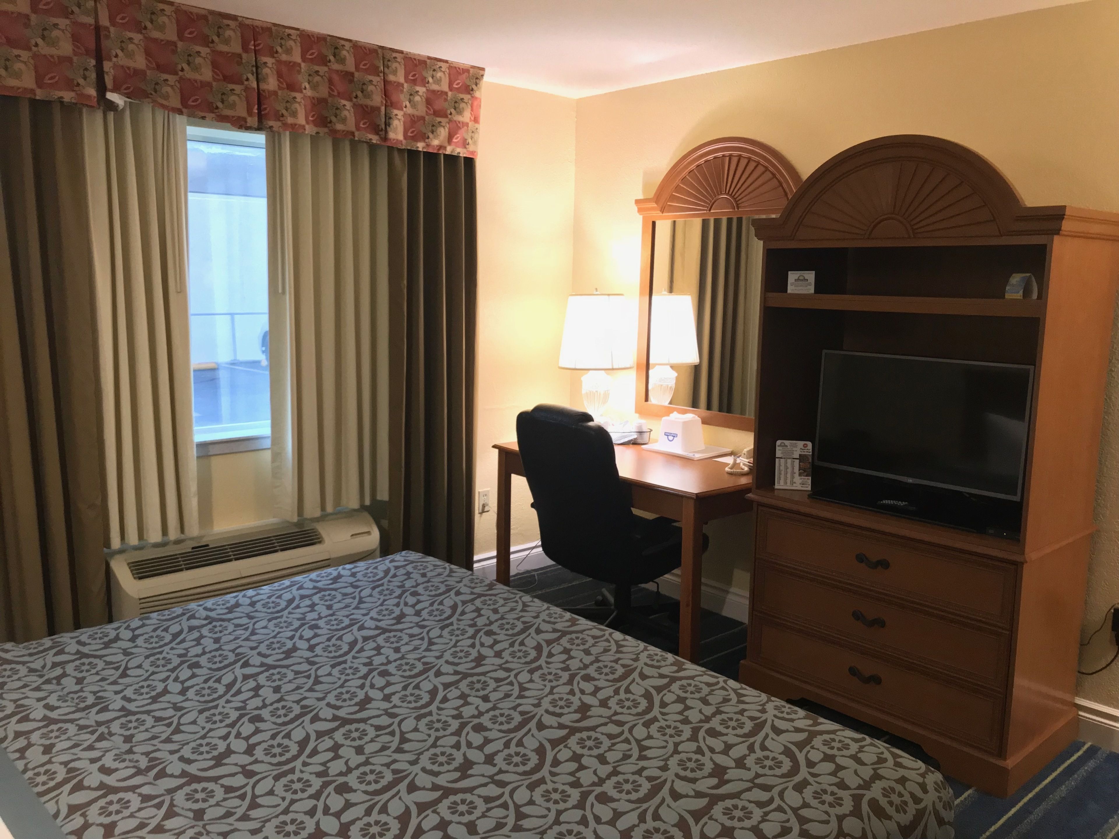 A hotel room with a bed, a desk with a chair, a television, a mirror, and a window with curtains.