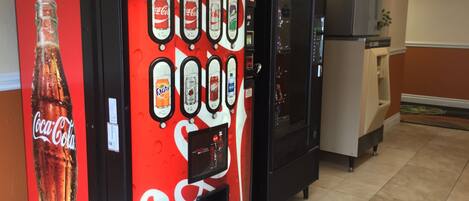 Máquina expendedora con diversas opciones de bebidas y un refrigerador en una habitación con suelo de baldosas.