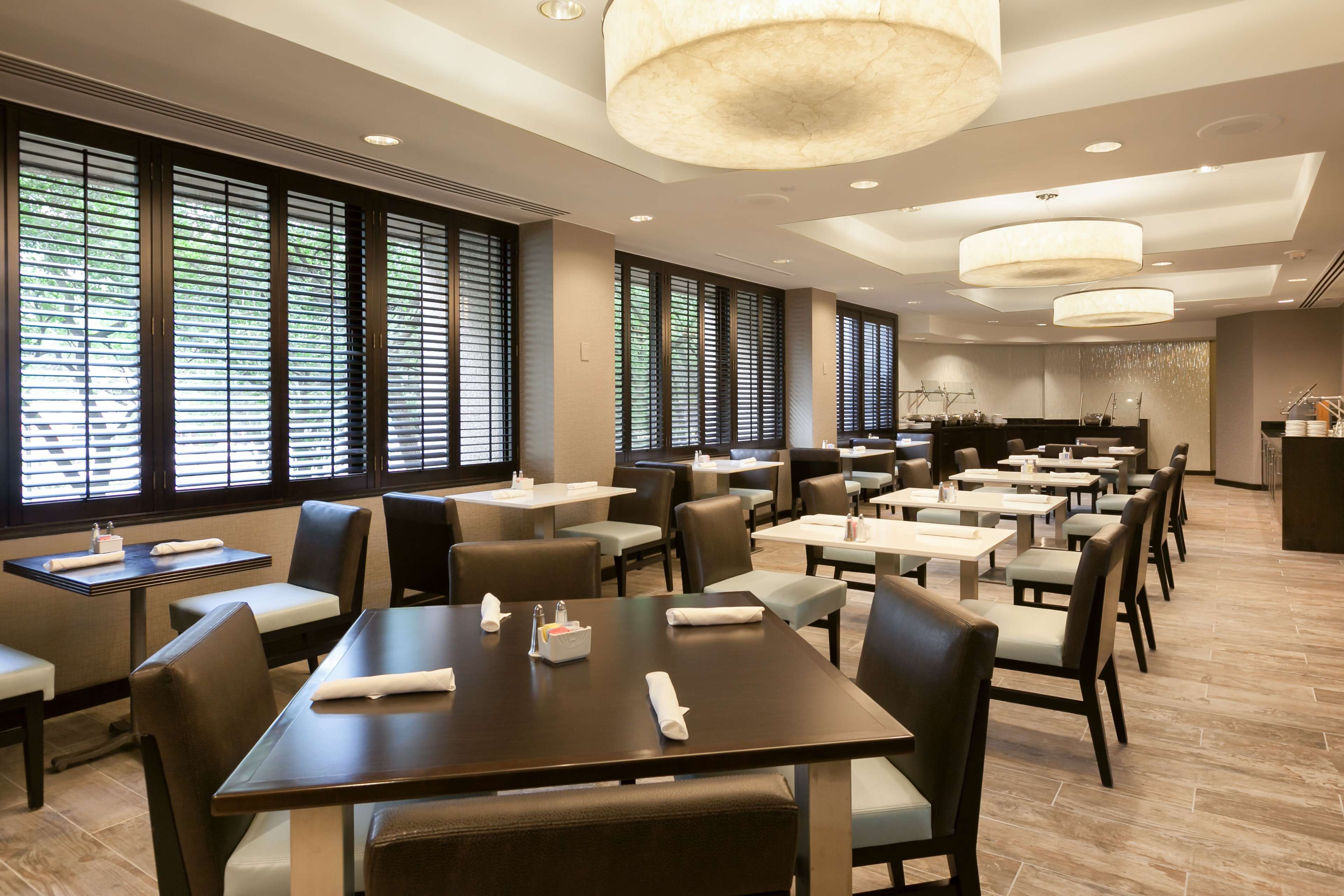 Un comedor amplio con varias mesas y sillas, ventanas grandes con persianas e iluminación colgante moderna.