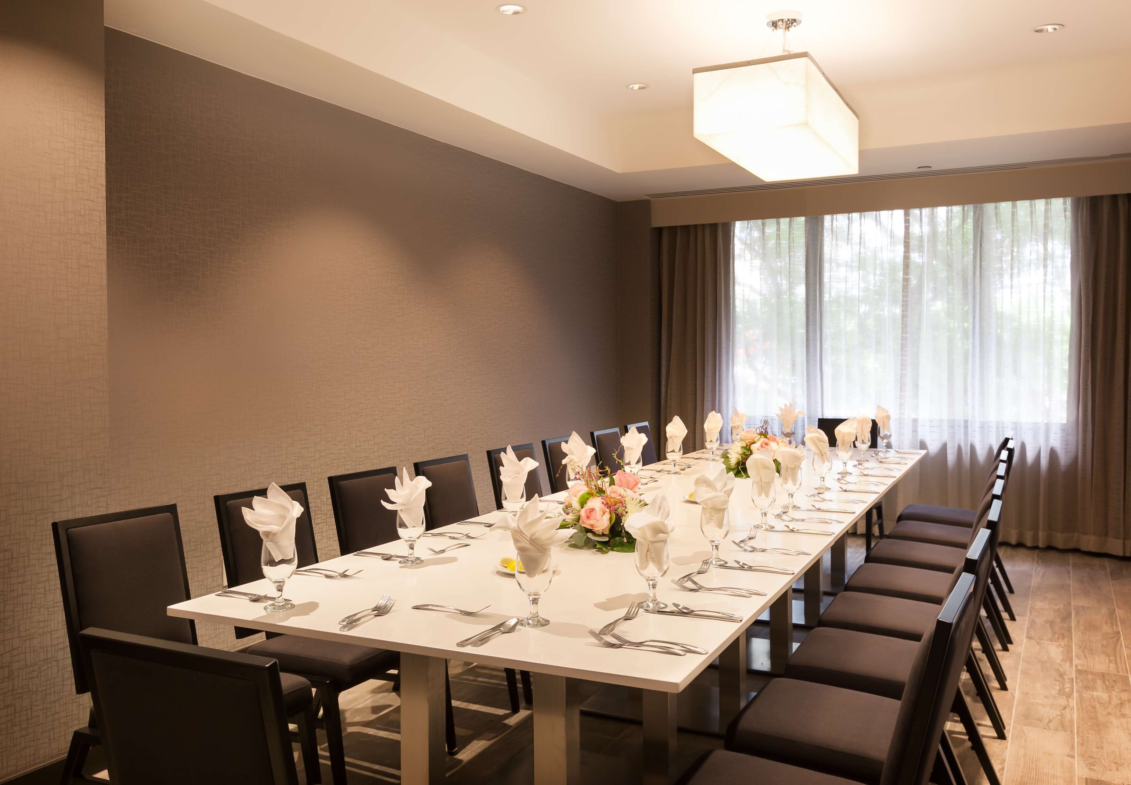 A long dining table set with white tablecloths, silverware, and floral centerpieces in a well-lit room with large windows and curtains.