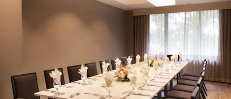 Una larga mesa de comedor vestida con manteles blancos, cubiertos y centros de mesa florales en una habitación luminosa con ventanas grandes y cortinas.