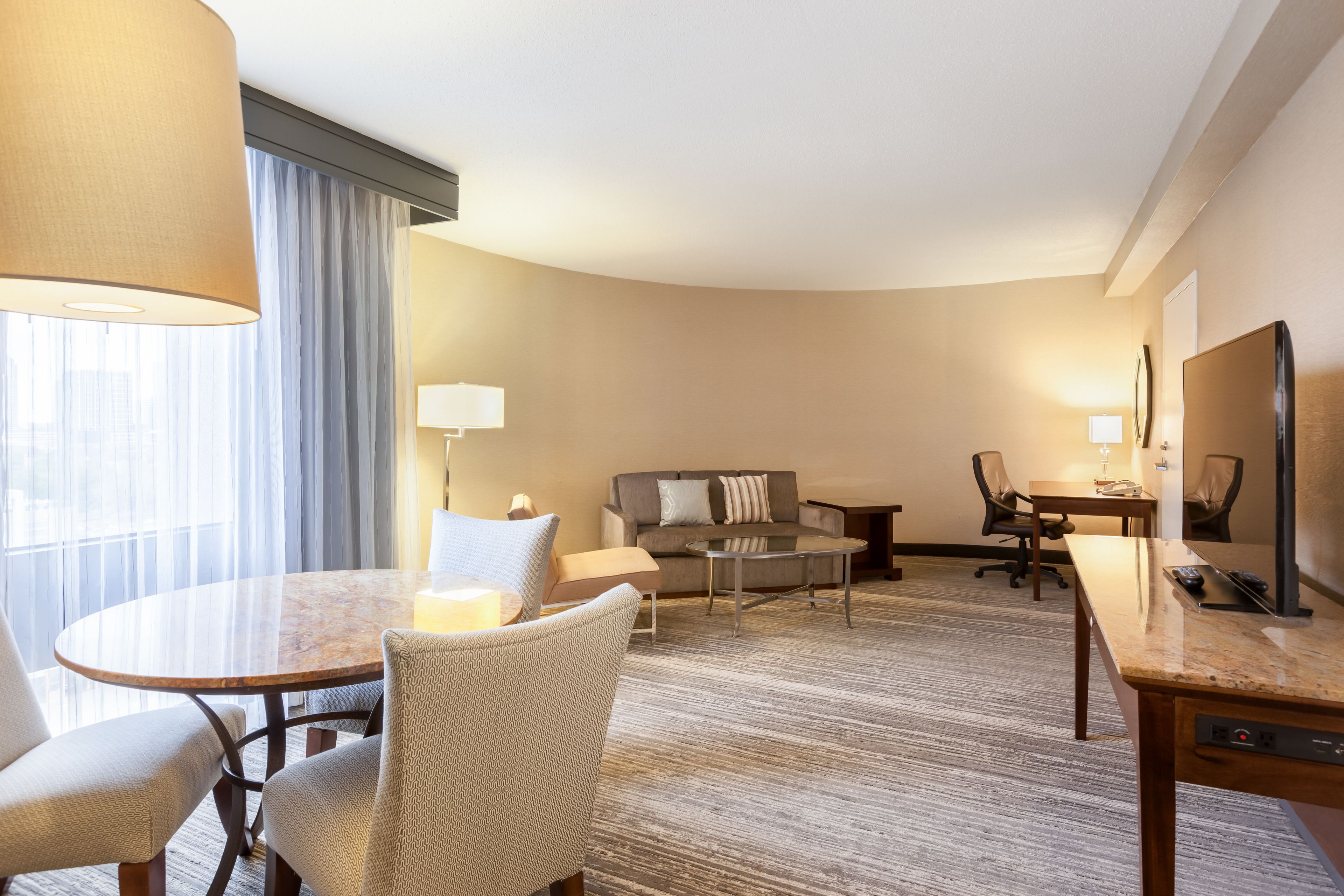 A hotel room with a round dining table, two armchairs, a sofa, a coffee table, a TV, and a large window with curtains.