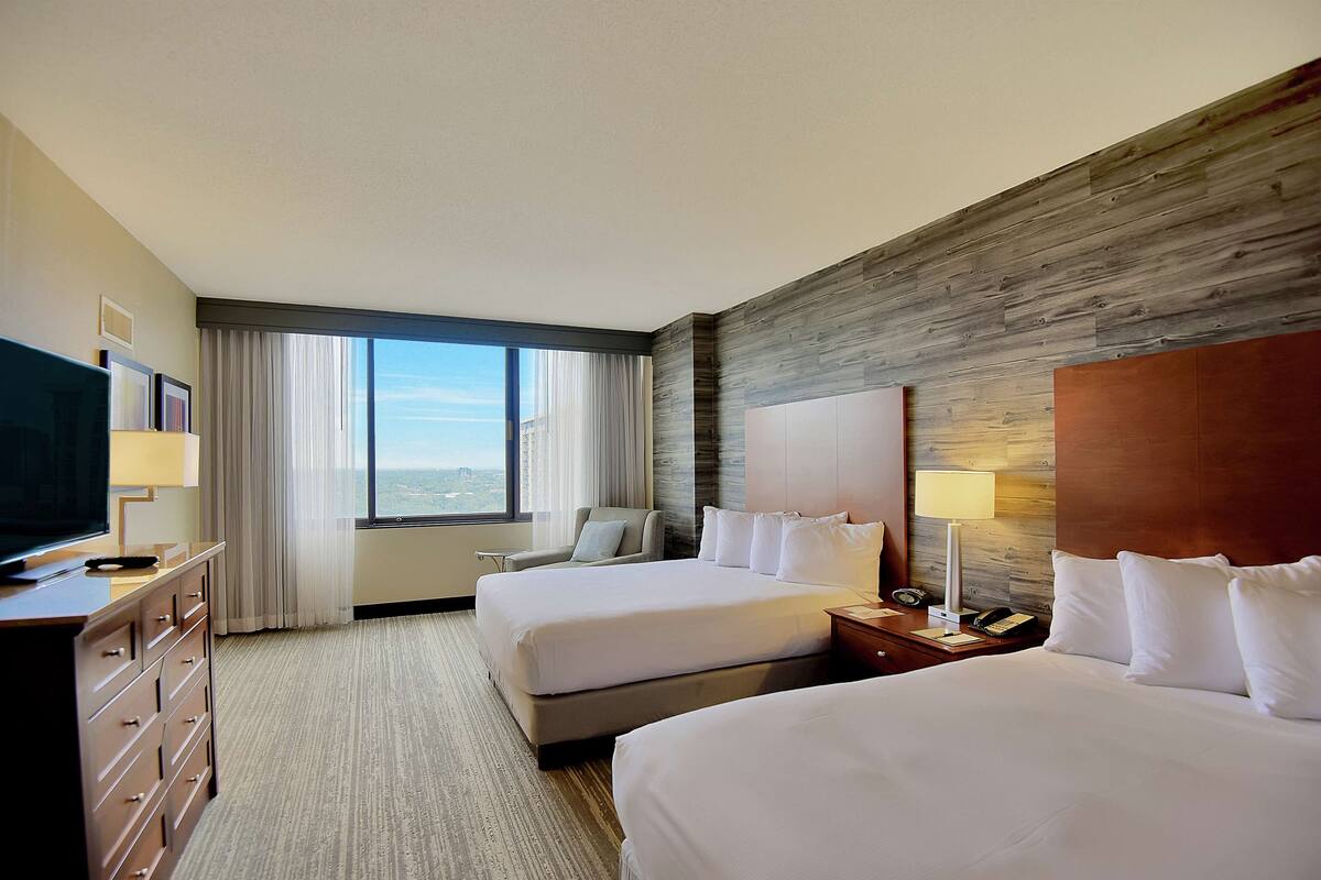Habitación de hotel con dos camas, televisor, escritorio y ventana grande con vistas.