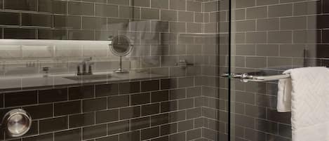 A modern bathroom with dark tiled walls, a glass shower enclosure, a white sink with a mirror, and a towel rack.