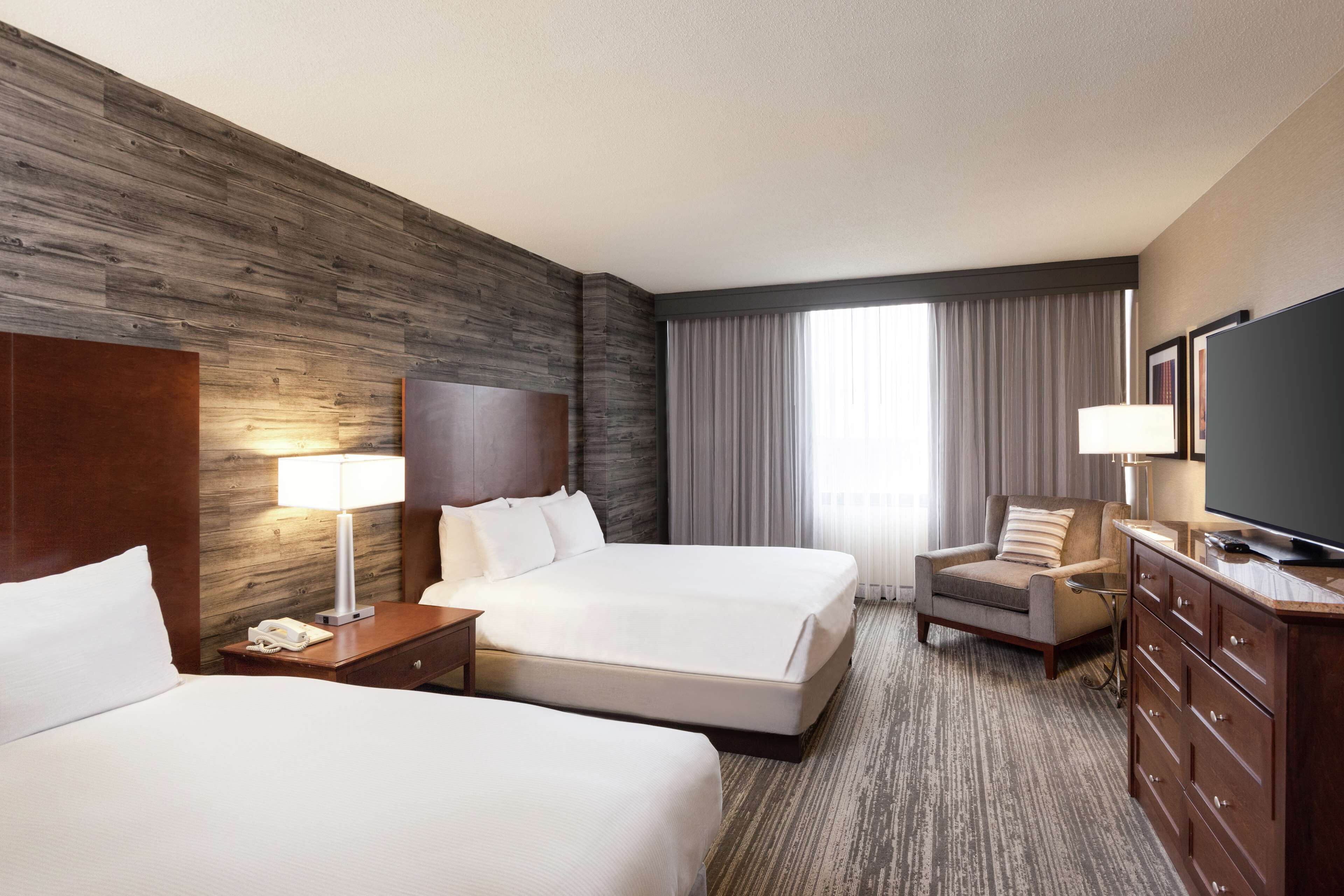 A hotel room with two beds, a desk, a chair, a TV, and a large window with curtains.