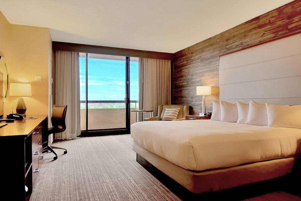 Habitación de hotel con una cama grande, un escritorio con lámpara, una silla y vistas al exterior a través de una puerta corrediza.