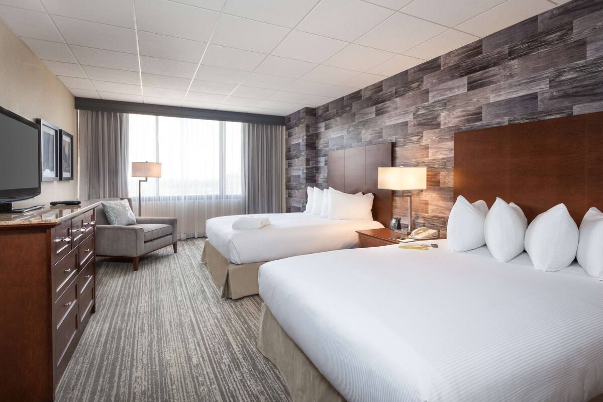 Habitación de hotel con dos camas, un escritorio, televisor y un ventanal grande con cortinas.