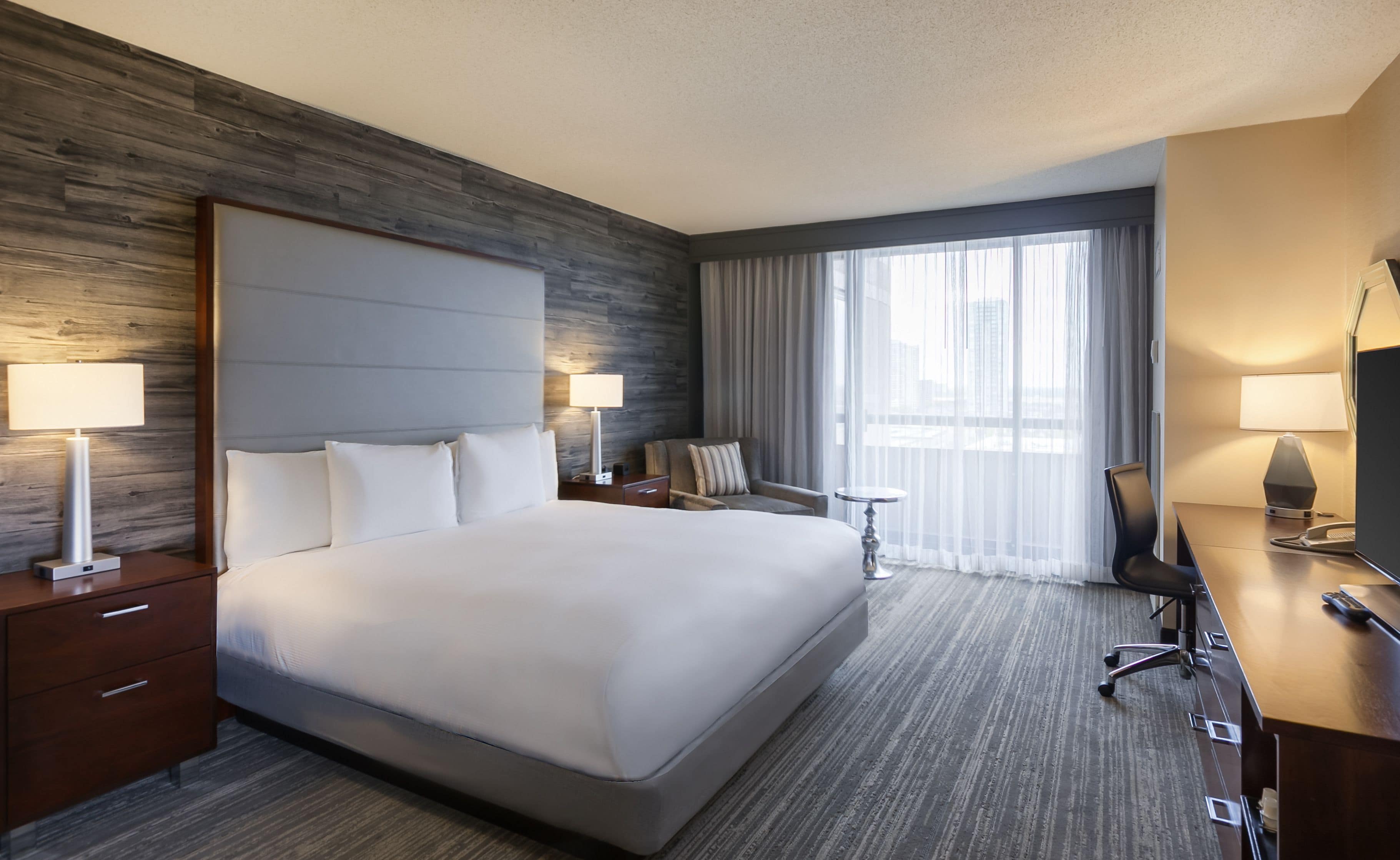 Une chambre d’hôtel avec un grand lit, un bureau, une chaise, une petite table et une fenêtre avec des rideaux.