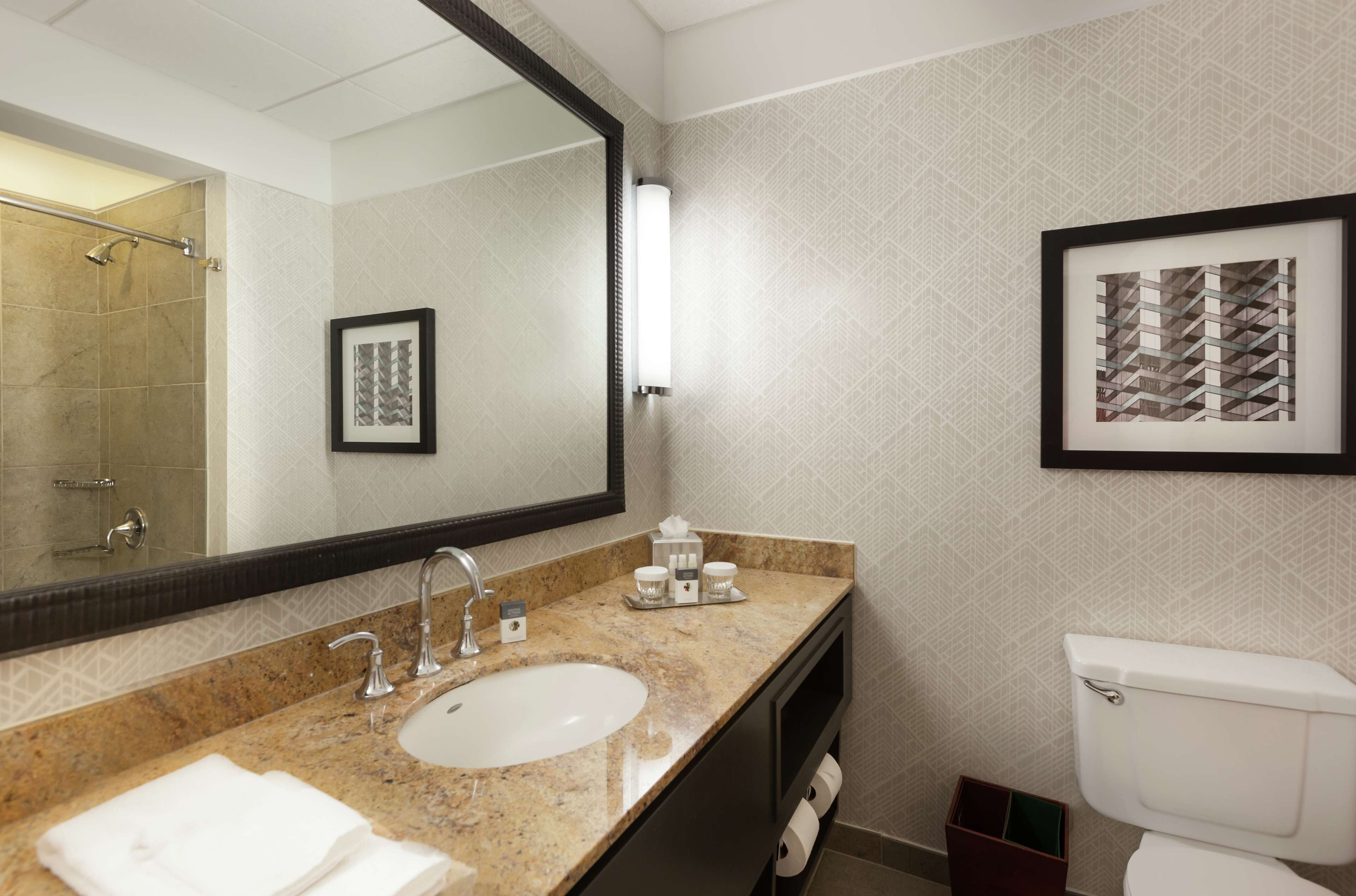 A bathroom with a large mirror, a sink, two faucets, a toilet, and a framed picture on the wall.