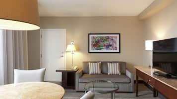 A hotel room with a round table, a sofa with pillows, a TV, a lamp, and a painting on the wall.