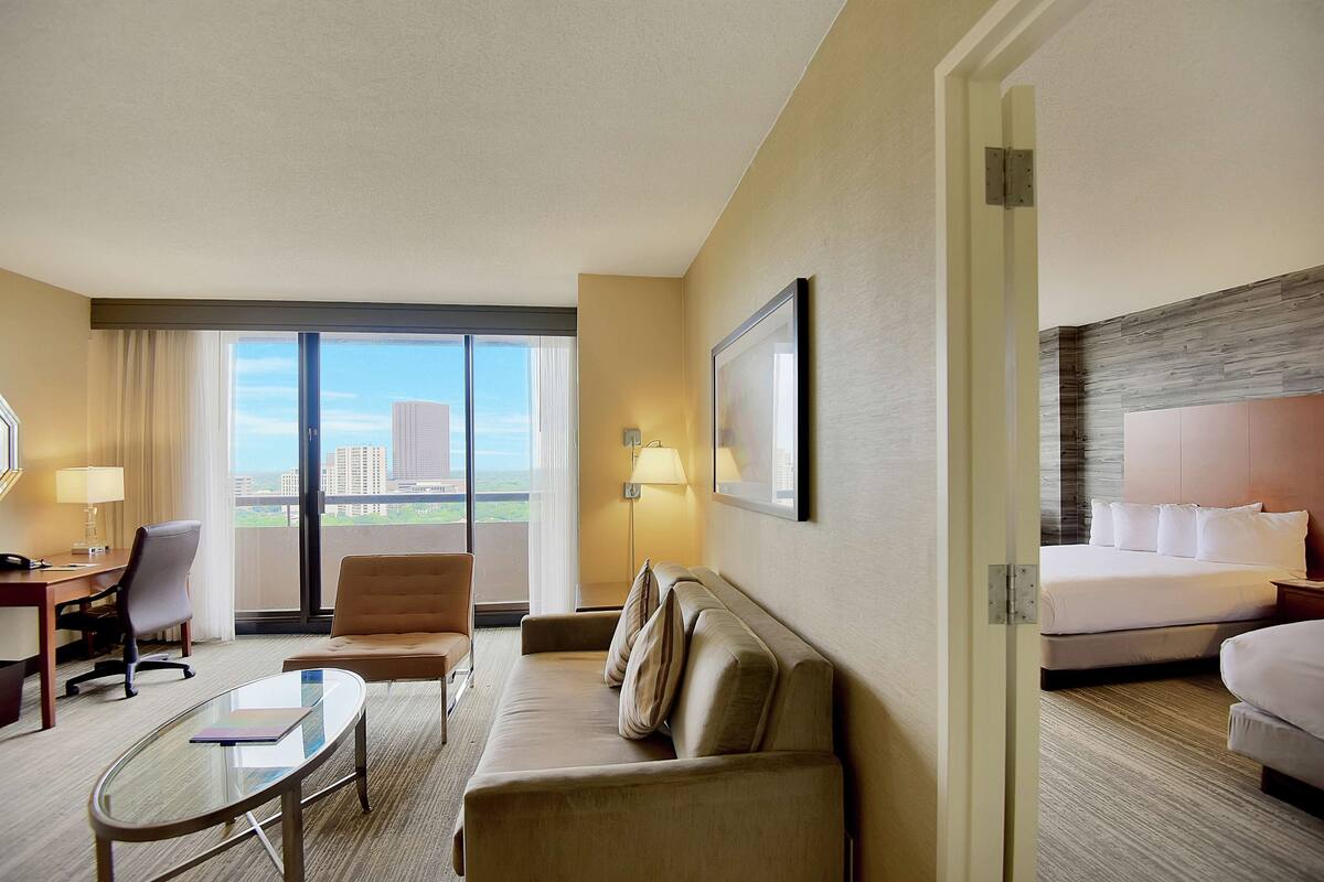 Habitación de hotel con un ventanal grande, un sofá, un escritorio con una silla, una cama y un cuadro en la pared.