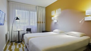 Une chambre d’hôtel avec deux lits, un bureau, une chaise, une lampe et un mur sur lequel est affiché un panneau indiquant « EN DIRECT ».