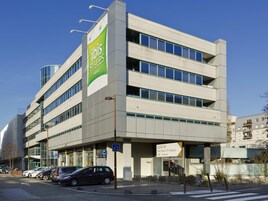 Un hotel de varios pisos con un llamativo letrero verde y blanco, numerosas ventanas y un estacionamiento con varios autos.
