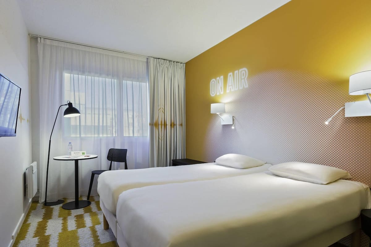 Ein Hotelzimmer mit zwei Betten, einem kleinen Tisch, einem Stuhl, einer Lampe und einer Wandleuchte.