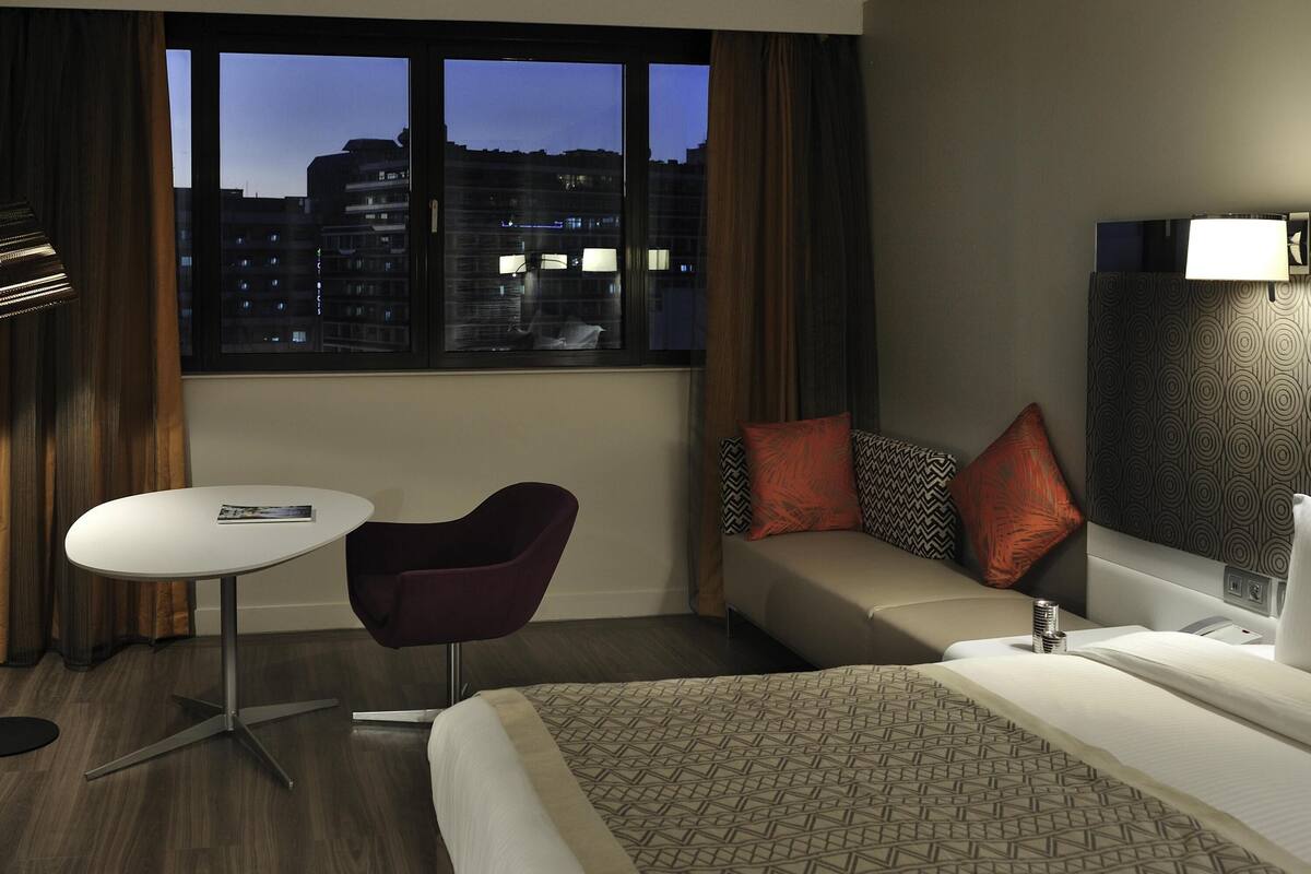 A hotel room with a bed, a sofa, a small table, and a chair. There is a window with a view of buildings outside.