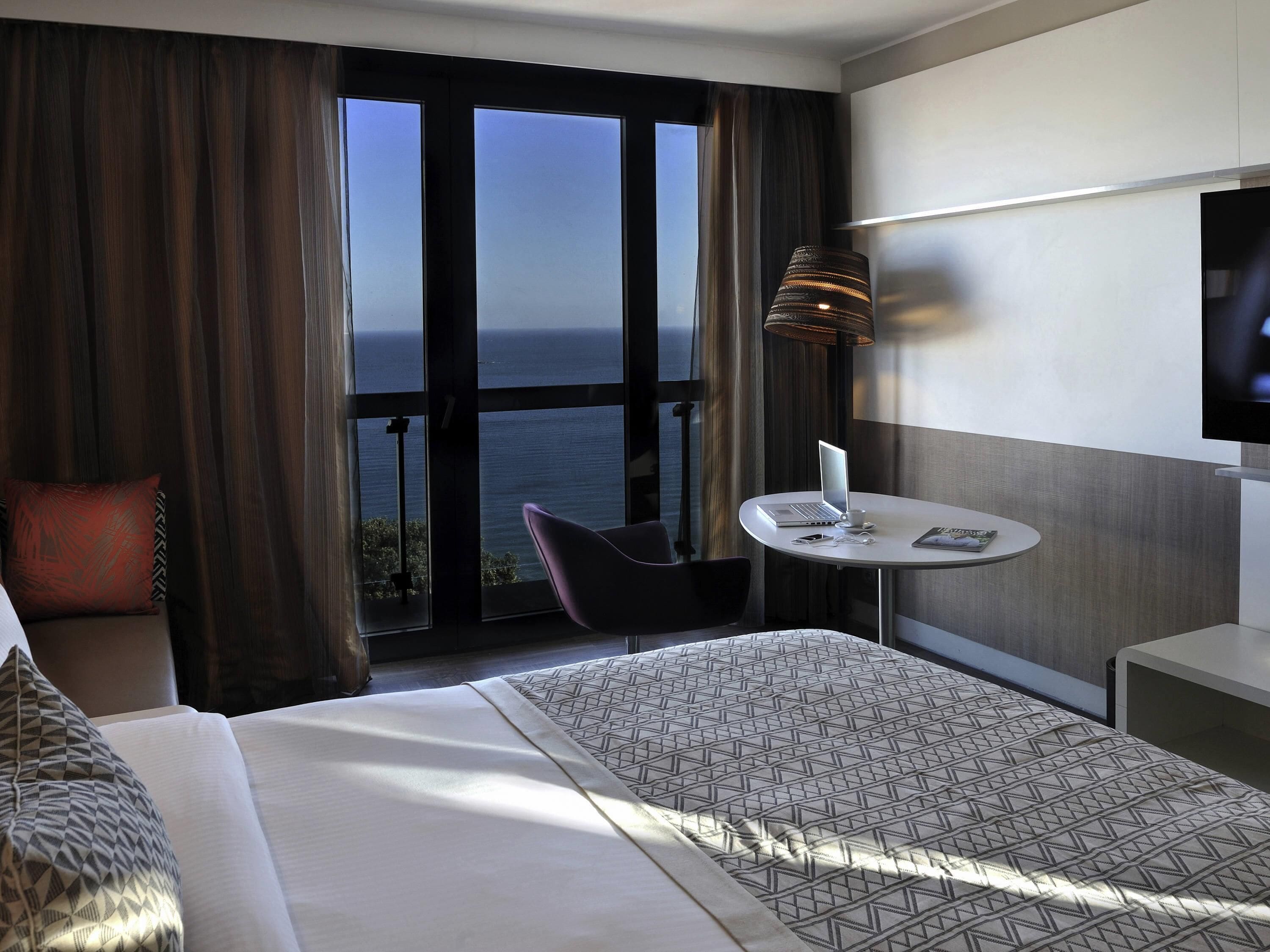 Habitación de hotel con cama, escritorio con computadora portátil, silla, televisor y vista al océano.