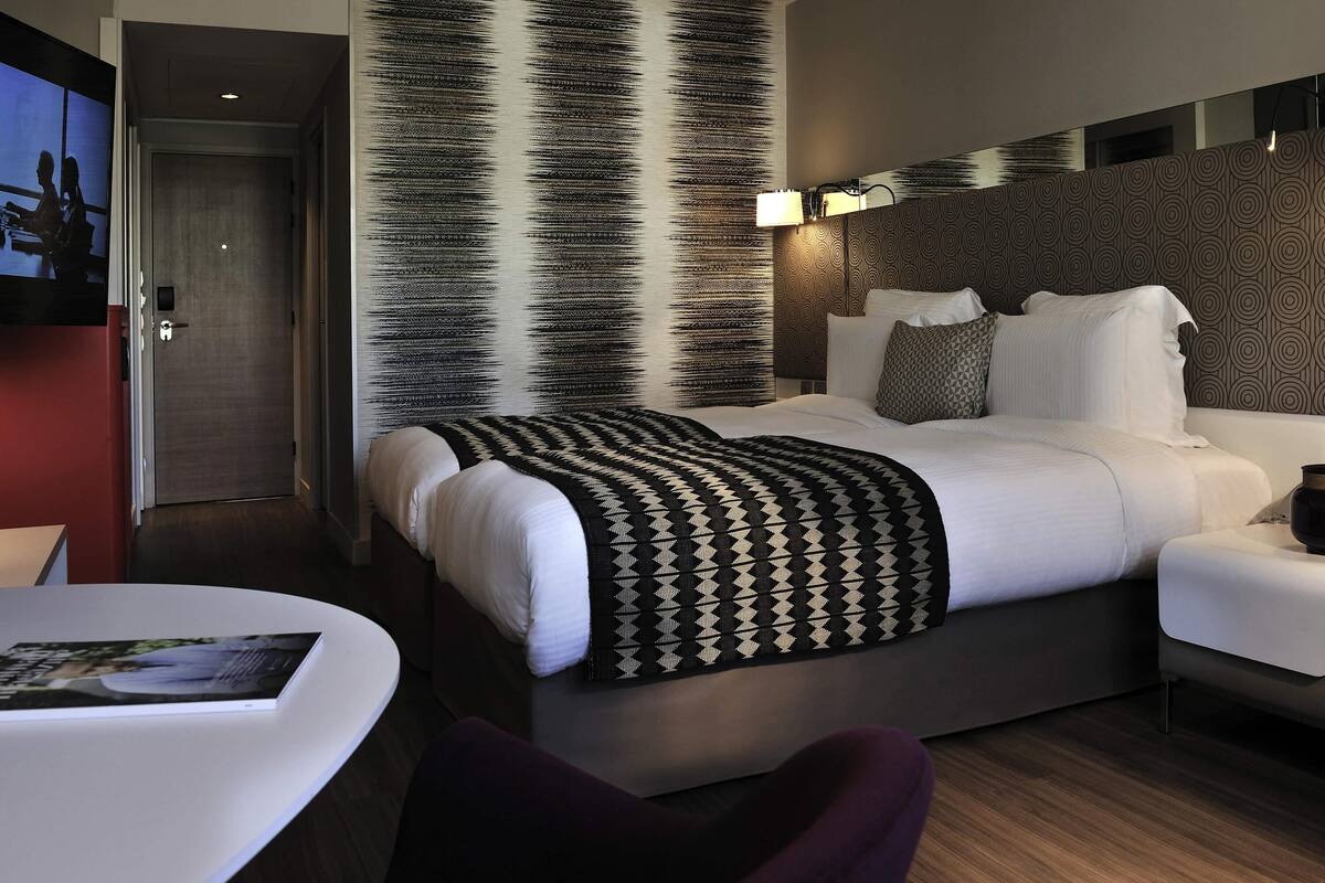 A modern hotel room with a large bed, a bedside table, a chair, and a wall-mounted TV.