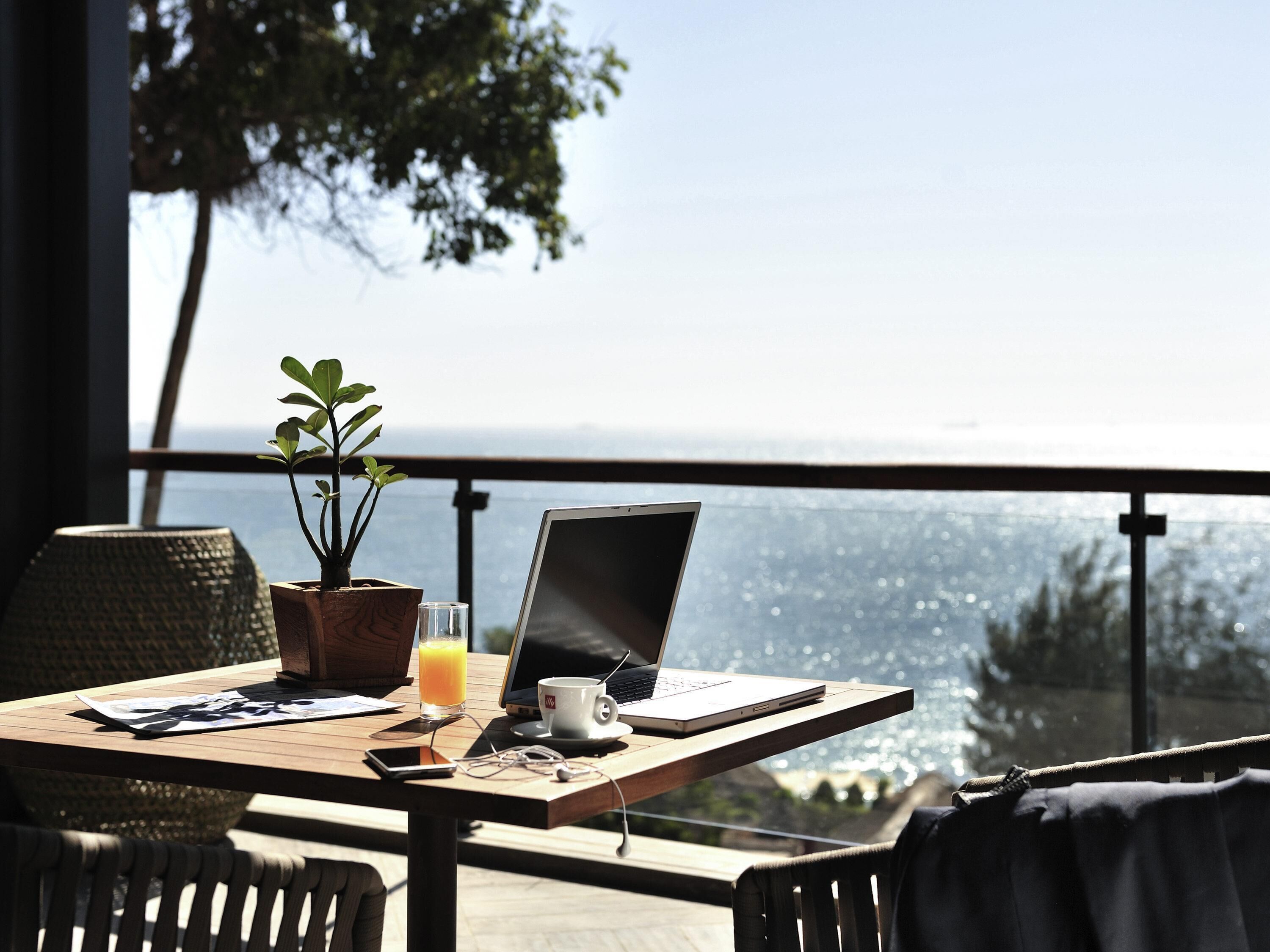 Ein Laptop auf einem Balkontisch mit einer Pflanze, einem Orangensaftglas und einer Tasse, mit Blick auf eine schöne Aussicht.