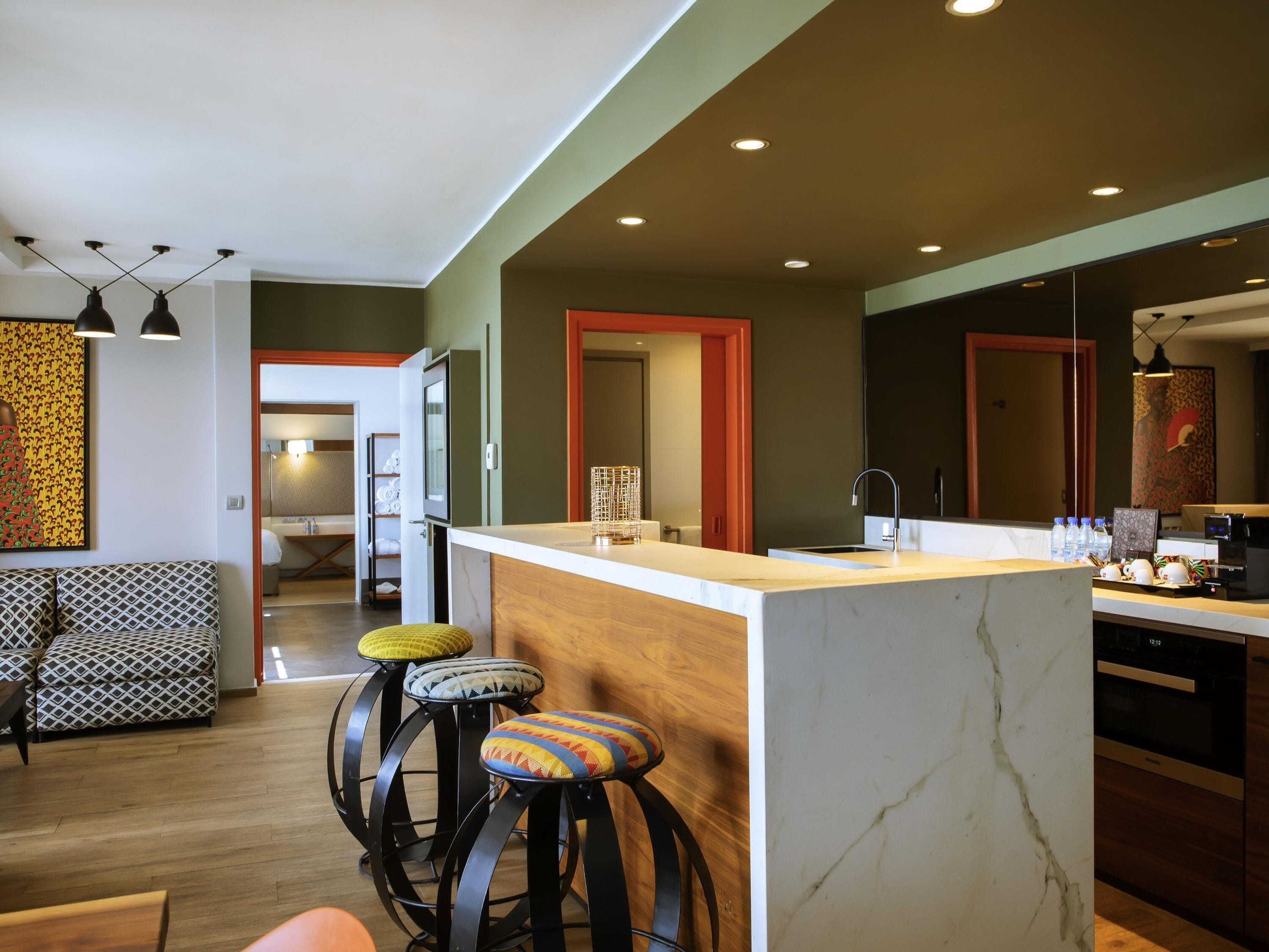A modern kitchen with a marble countertop, a sink, and a bar area with stools. There is a wooden dining table and a patterned sofa in the adjacent living area.