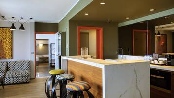 A modern kitchen with a marble countertop, a sink, and a bar area with stools. There is a wooden dining table and a patterned sofa in the adjacent living area.